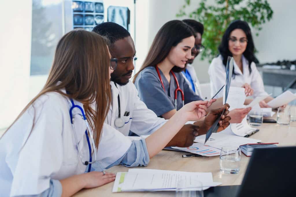 Group Of Doctors Are Studying Disease Patient's History. Team Of Multiethnic Young Doctors Having A Meeting In Conference Room In The Modern Bright Hospital.