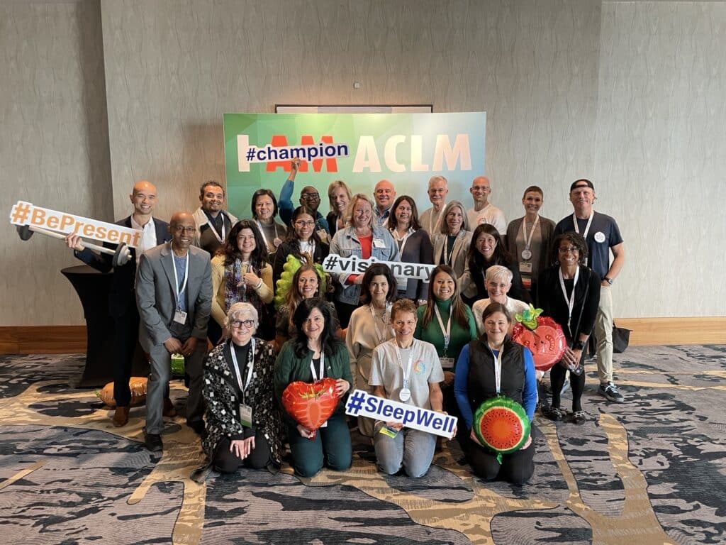 Group of people standing in front of a photo backdrop saying "I AM ACLM" holding up motivational signs and balloons that are shaped like fruit