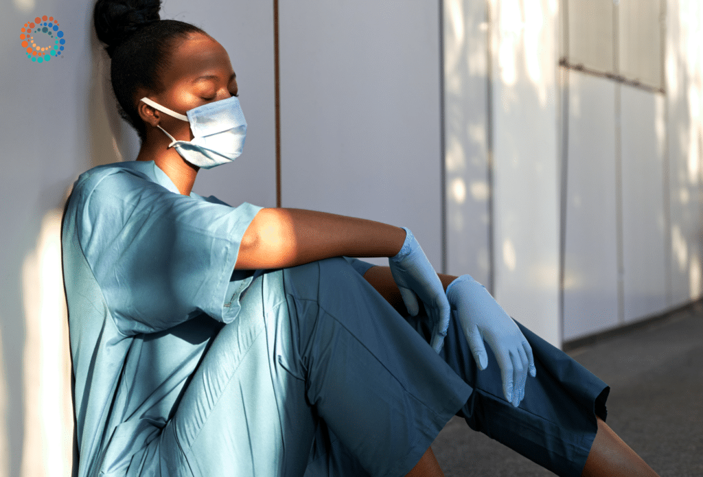 Healthcare professional sitting on the ground in a hallway looking down with a mask on exhibiting the sense of burnout